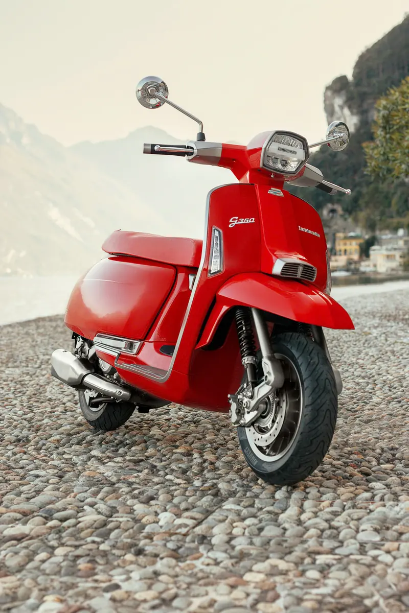 Lambretta G350 màu đỏ trên bờ hồ lát sỏi, phía sau là núi